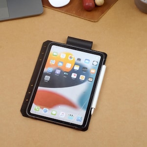 Może przedstawiać: Czarny tablet z dołączonym białym rysikiem, spoczywający na brązowej powierzchni. Tablet wyświetla kolorowy ekran aplikacji. Tablet jest w czarnym etui z klipsem na g&oacute;rze.