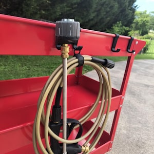 May include: A red utility cart with a pressure washer nozzle and hose. The nozzle is black and gray, attached to a brass fitting. The hose is tan and coiled. The cart has black hooks and red wheels.