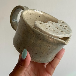 May include: A speckled beige ceramic tea strainer mug with a curved handle and a built-in perforated lid section over the spout. The glaze has a glossy finish with dark brown flecks throughout the surface, featuring a wide opening and a small pouring spout.