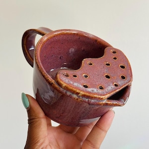 Puede incluir: Una taza de cerámica color magenta intenso con un borde colador integrado en la parte superior. El esmalte brillante y moteado cubre el exterior y el interior, mientras que la sección del colador contiene múltiples orificios circulares.