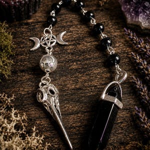 May include: A pendulum featuring a series of smooth black beads connected to a silver-toned metal charm of a pentagram flanked by two crescent moons. Below this hangs a silver-toned bird skull charm and a faceted black stone point pendant.