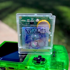 Op de afbeelding: Een doorzichtige plastic videogamecartridge met een holografisch label met de tekst Pokémon Gotta catch em all! en een afbeelding van Gengar, rustend op een doorschijnende neon-groene handheld spelcomputer met een zwart richtingspad.