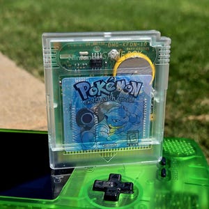 May include: A transparent Game Boy video game cartridge for Pokémon Blue Version featuring a blue Blastoise illustration and the text Pokémon Gotta catch 'em all! resting on a translucent neon green handheld video game console device.
