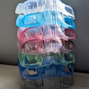 Puede incluir: Una pila de seis clips de plástico transparente en varios colores: transparente, azul, rosa, verde y gris. Cada clip tiene una forma curva con una parte superior con bisagras y una abertura rectangular. Los clips están apilados.