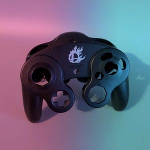 Puede incluir: Un mando de GameCube negro con un logotipo blanco de Super Smash Bros. y las palabras "Start Pause" impresas en la parte delantera.