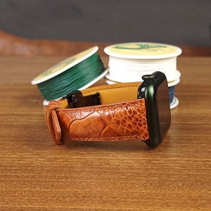 May include: A brown leather watch band with a textured pattern. The band is attached to a black and green smartwatch.