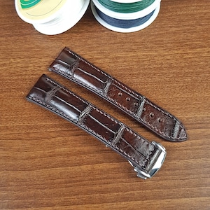 May include: Two dark brown leather watch straps with a crocodile skin pattern. The straps are angled, showing the buckle and the holes for the watch pins. Spools of thread are in the background.