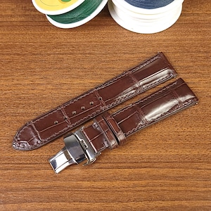 May include: A brown leather watch strap with a silver buckle. The strap has a crocodile pattern.