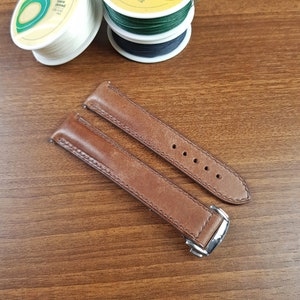 May include: Two brown leather watch straps with stitching detail. The straps have a silver buckle and are laid out on a wooden surface.