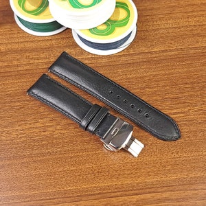 Könnte beinhalten: Zwei schwarze Lederarmbänder mit einer silbernen Schließe. Die Armbänder liegen auf einer Holzoberfläche.