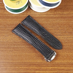 May include: Two black watch straps with a textured, ribbed design. The straps have a silver-colored clasp and are lying on a wooden surface. Spools of thread in various colors are visible in the background.