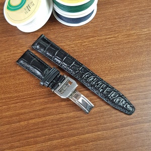 Può includere: Cinturino da orologio in pelle di alligatore nero con fibbia argentata. Il cinturino è disposto su una superficie in legno.