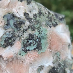 Op de afbeelding: Close-up van een mineraalmonster met een mix van kleuren en texturen. De steen toont een combinatie van witte, perzik, zwarte en groene tinten. De kristalformaties hebben een radiaal patroon.