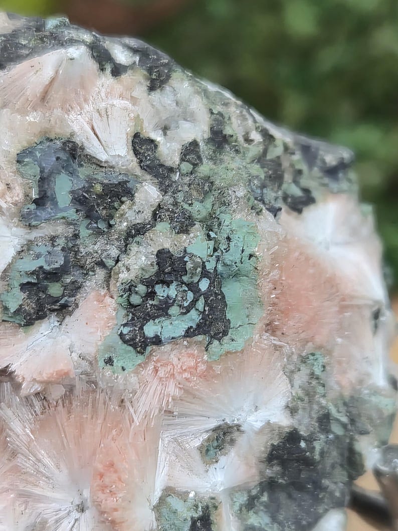 Op de afbeelding: Close-up van een mineraalmonster met een mix van kleuren en texturen. De steen toont een combinatie van witte, perzik, zwarte en groene tinten. De kristalformaties hebben een radiaal patroon.