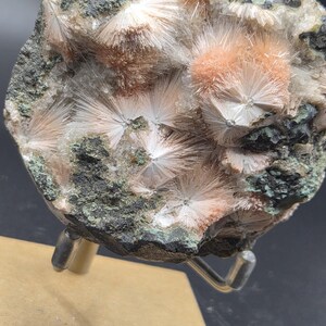 Op de afbeelding: Een close-up van een mineraalmonster met clusters van uitstralende, naaldachtige kristallen in perzik- en wittinten. De kristallen zijn geplaatst tegen een donkere, gespikkelde matrix met groene vlekken. Het monster is gemonteerd op een metalen standaard.