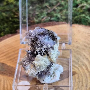 May include: A mineral specimen encased in a clear acrylic display box. The mineral features a mix of colors, including purple, white, and a touch of green. The box is open, revealing the intricate details of the natural formation.