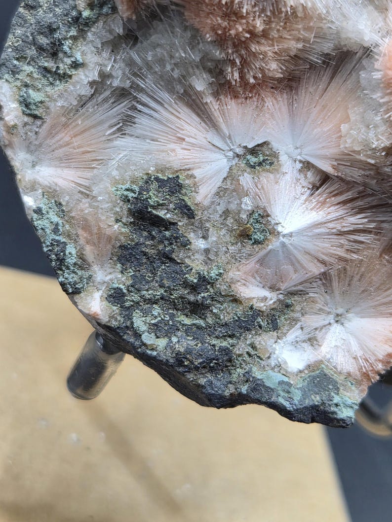 Op de afbeelding: Close-up van een mineraalmonster met stralende clusters van witte, naaldachtige kristallen. De kristallen zijn geplaatst tegen een achtergrond van donkergrijze en groene rotsen, met hints van perzikgekleurde formaties.