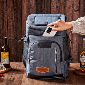 Puede incluir: Una mochila gris aislante con detalles negros y cremallera arco&iacute;ris. Un tel&eacute;fono est&aacute; en el bolsillo delantero. La mochila tiene el texto "HERMAN GROOMSMAN" en un parche de cuero.