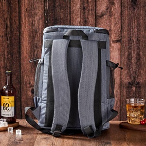 Puede incluir: Una mochila nevera gris con correas negras y bolsillos laterales de malla. Dos botellas de cerveza con etiquetas coloridas y cubitos de hielo est&aacute;n sobre una superficie de madera. Tambi&eacute;n hay un vaso de t&eacute; helado y cerezas.