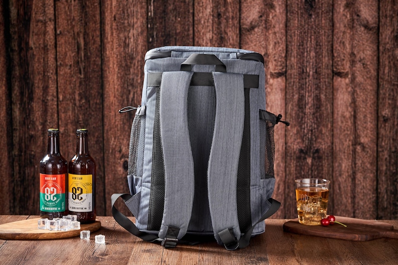 Puede incluir: Una mochila nevera gris con correas negras y bolsillos laterales de malla. Dos botellas de cerveza con etiquetas coloridas y cubitos de hielo est&aacute;n sobre una superficie de madera. Tambi&eacute;n hay un vaso de t&eacute; helado y cerezas.