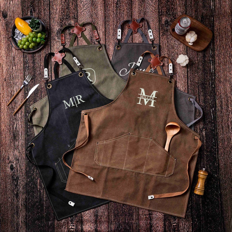 May include: A collection of personalized aprons in various colors, including black, brown, and olive green. Each apron features a pocket and leather straps, with embroidered initials or names. A wooden spoon rests in one pocket.