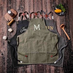 May include: Three canvas aprons with leather accents and personalized embroidery. The aprons are in shades of black, olive green, and gray. Each apron has a large front pocket and adjustable straps. The aprons are displayed on a wooden surface.