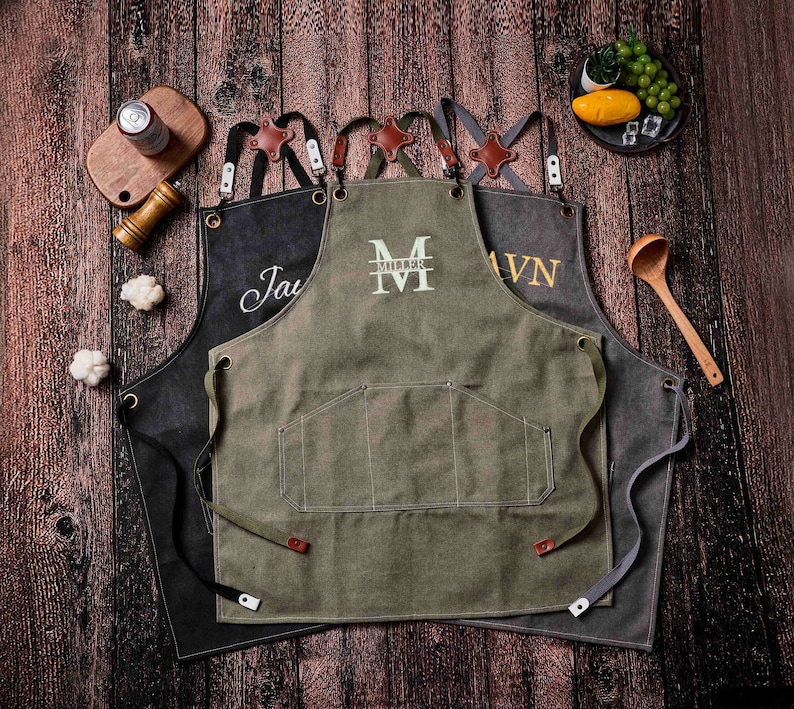 May include: Three canvas aprons with leather accents and personalized embroidery. The aprons are in shades of black, olive green, and gray. Each apron has a large front pocket and adjustable straps. The aprons are displayed on a wooden surface.