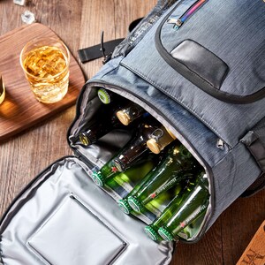 Puede incluir: Una bolsa nevera gris con cremallera arco&iacute;ris y asa negra est&aacute; abierta, revelando varias botellas de cerveza verdes. Un vaso de l&iacute;quido &aacute;mbar y cubitos de hielo est&aacute;n sobre una superficie de madera.
