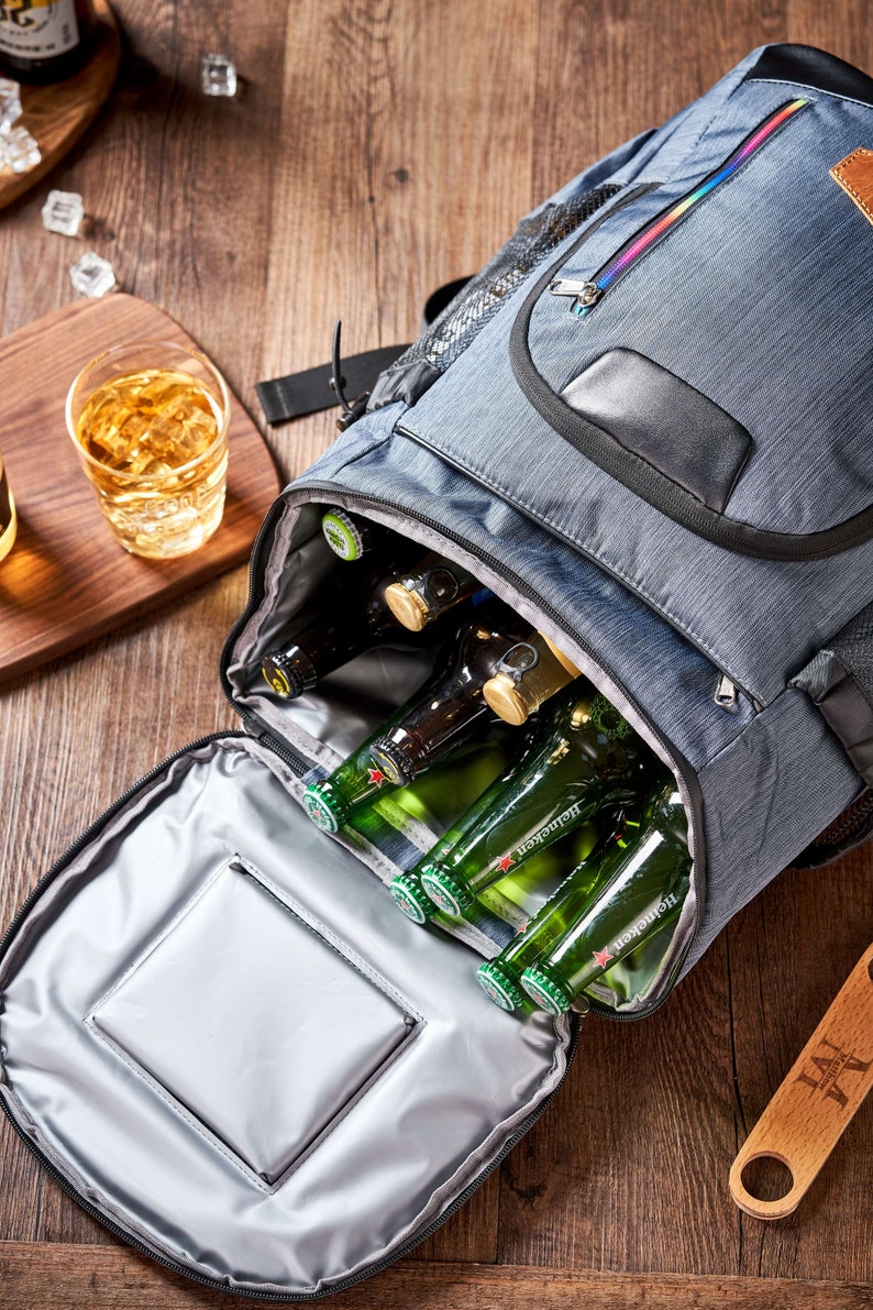 Puede incluir: Una bolsa nevera gris con cremallera arco&iacute;ris y asa negra est&aacute; abierta, revelando varias botellas de cerveza verdes. Un vaso de l&iacute;quido &aacute;mbar y cubitos de hielo est&aacute;n sobre una superficie de madera.