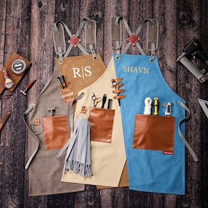 May include: Three personalized aprons in shades of gray, tan, and blue, with brown leather pockets and straps. The aprons have the initials "R/S" and the name "SHAVN" embroidered on them. Various barber tools are visible.