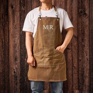 May include: Brown canvas apron with a large front pocket and leather straps. The apron has the embroidered initials "MR" in white. The apron is worn by a person wearing a white t-shirt and blue jeans.