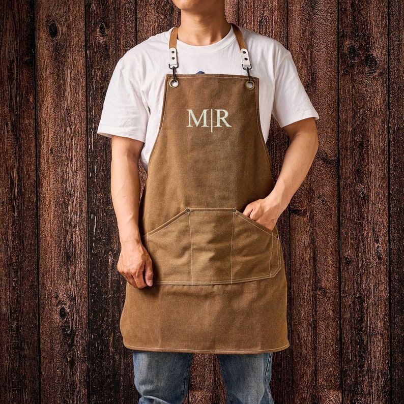 May include: Brown canvas apron with a large front pocket and leather straps. The apron has the embroidered initials "MR" in white. The apron is worn by a person wearing a white t-shirt and blue jeans.