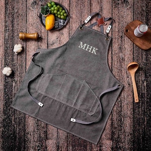 May include: Charcoal gray apron with the embroidered initials "MHK". The apron has a large front pocket and adjustable straps. The apron is made of a durable material and is designed for cooking or grilling.
