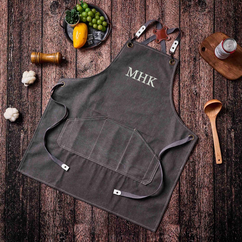 May include: Charcoal gray apron with the embroidered initials "MHK". The apron has a large front pocket and adjustable straps. The apron is made of a durable material and is designed for cooking or grilling.