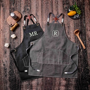 May include: Two aprons with leather straps and pockets are displayed on a wooden surface. One is black with "MR" embroidered in white, the other is gray with an "R" in a white circle. The aprons have brown leather accents.