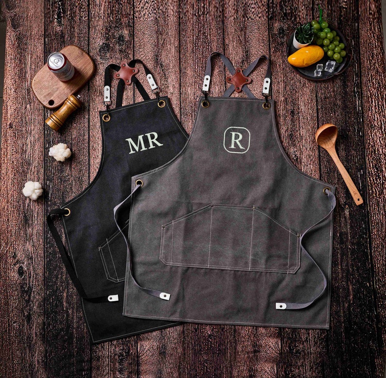 May include: Two aprons with leather straps and pockets are displayed on a wooden surface. One is black with "MR" embroidered in white, the other is gray with an "R" in a white circle. The aprons have brown leather accents.