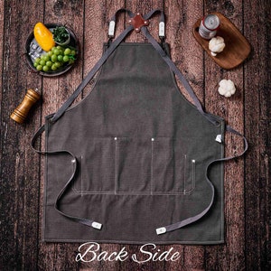 May include: Charcoal gray apron with three front pockets and adjustable straps. The apron has a leather and metal detail at the top and the text "Back Side" at the bottom. The apron is displayed on a wooden surface.