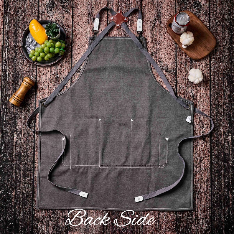 May include: Charcoal gray apron with three front pockets and adjustable straps. The apron has a leather and metal detail at the top and the text "Back Side" at the bottom. The apron is displayed on a wooden surface.