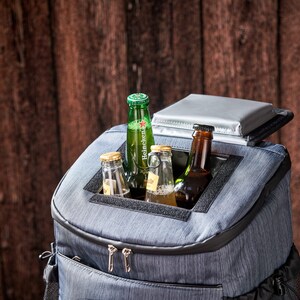 Puede incluir: Una nevera tipo mochila azul aislante con un bolsillo frontal con cremallera y un compartimento superior que contiene varias botellas de cerveza de vidrio. La nevera tiene una tapa plateada y un ribete negro, sobre un fondo de madera.