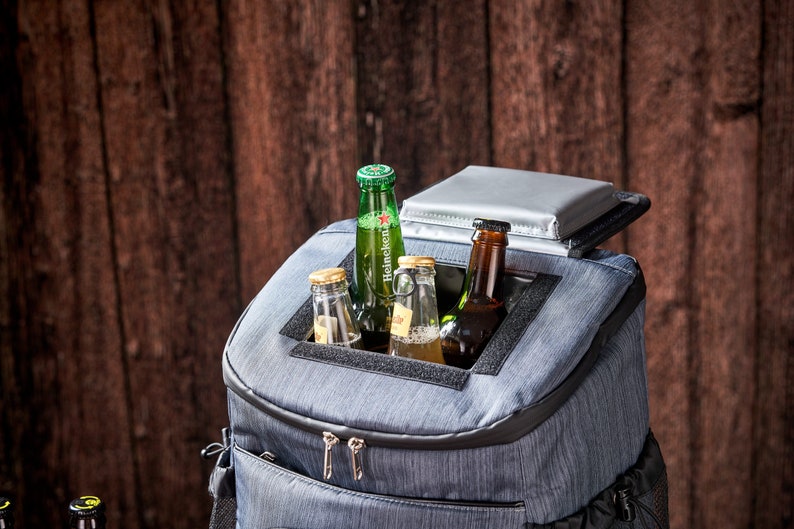 Puede incluir: Una nevera tipo mochila azul aislante con un bolsillo frontal con cremallera y un compartimento superior que contiene varias botellas de cerveza de vidrio. La nevera tiene una tapa plateada y un ribete negro, sobre un fondo de madera.