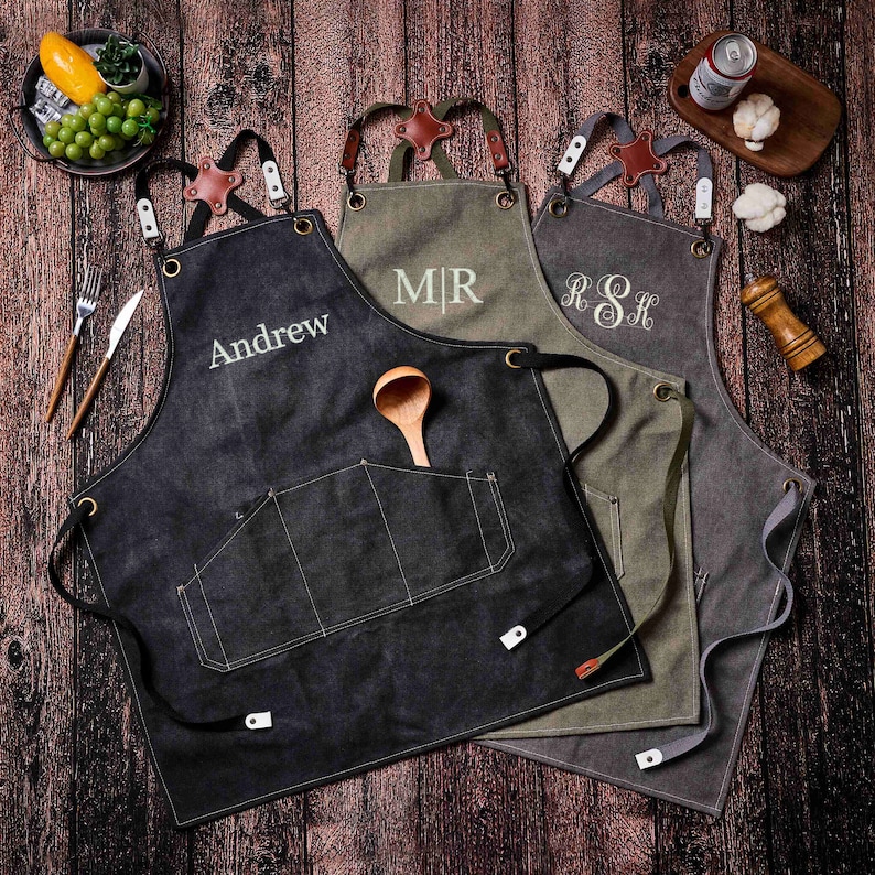 May include: Four personalized aprons in black, olive green, and gray, with leather and metal accents. Each apron features a pocket and is embroidered with a name or initials. A wooden spoon rests in the pocket of the black apron.