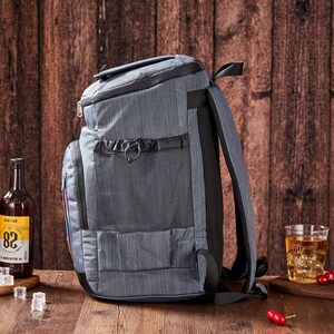 Puede incluir: Una mochila gris aislante con m&uacute;ltiples compartimentos y un asa negra. La mochila est&aacute; junto a varias botellas de cerveza y un vaso de hielo. El fondo es una pared de madera.