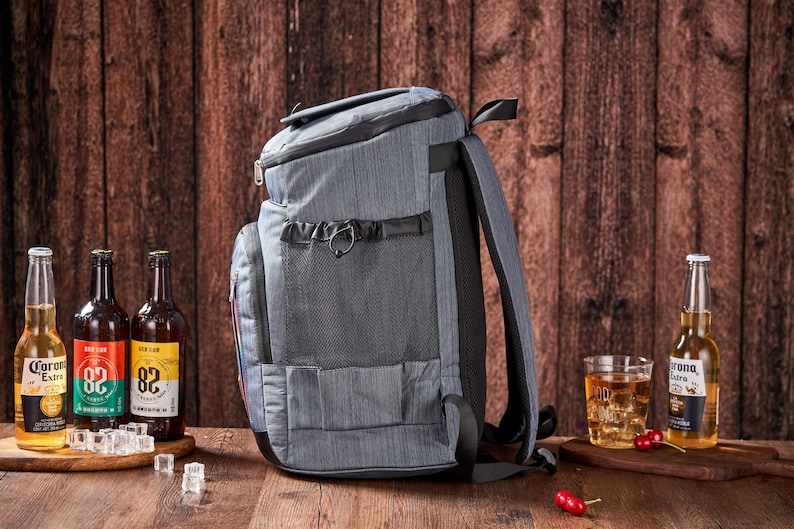Puede incluir: Una mochila gris aislante con m&uacute;ltiples compartimentos y un asa negra. La mochila est&aacute; junto a varias botellas de cerveza y un vaso de hielo. El fondo es una pared de madera.