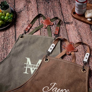 May include: Two canvas aprons, one olive green and one brown, with leather and metal accents. The olive green apron has the text "MILLER IV" embroidered on it. The brown apron has the text "Jauce" embroidered on it.