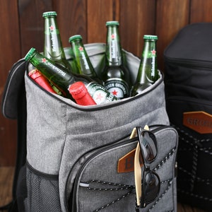 Pu&ograve; includere: Un zaino termico grigio riempito di bottiglie di birra verdi e bottiglie con tappo rosso. Occhiali da sole neri sono agganciati alla tasca anteriore. Una borsa termica nera &egrave; sullo sfondo. Le borse termiche sono per attivit&agrave; all'aperto.