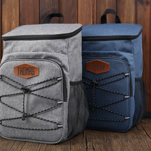 Pu&ograve; includere: Due zaini isolanti, uno grigio e uno blu, con dettagli neri. Ogni zaino presenta una toppa in pelle marrone con testo personalizzato. Lo zaino grigio ha il nome "THOMAS" e quello blu le iniziali "AKH".