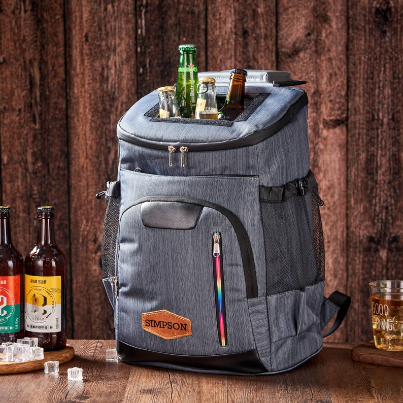 Puede incluir: Una nevera tipo mochila aislante gris con base y ribetes negros. La nevera tiene una cremallera arco&iacute;ris y un parche de cuero marr&oacute;n que dice "SIMPSON". Varias botellas de cerveza son visibles dentro de la nevera y sobre la mesa.