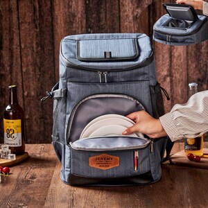 Puede incluir: Una mochila nevera gris con m&uacute;ltiples compartimentos, incluyendo una secci&oacute;n superior para enfriar y un bolsillo frontal. El bolsillo frontal contiene platos blancos. Un parche de cuero dice "JEREMY GROOMSMAN 05.22.2023".