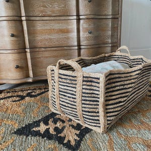 Puede incluir: Una cesta rectangular de yute con rayas horizontales negras y dos asas. La cesta está colocada sobre una alfombra estampada, con una cómoda de madera en el fondo. La cesta contiene una manta doblada.