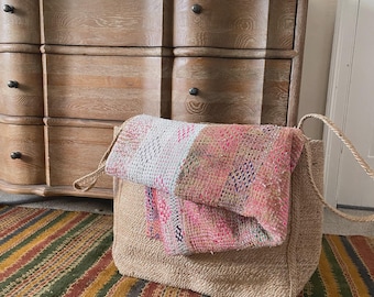 Puede incluir: Un bolso tote de yute tejido con asas de cuerda, lleno de un textil estampado colorido. El bolso está sobre una alfombra a rayas con tonos verdes, amarillos y rojos. Un aparador de madera se encuentra en el fondo.